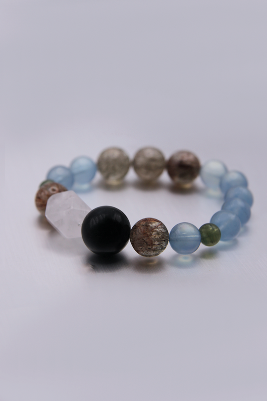 A beaded stretch bracelet featuring a variety of polished and textured beads in light blue, white, black, brown, and green, arranged in a circular shape on a light gray background.